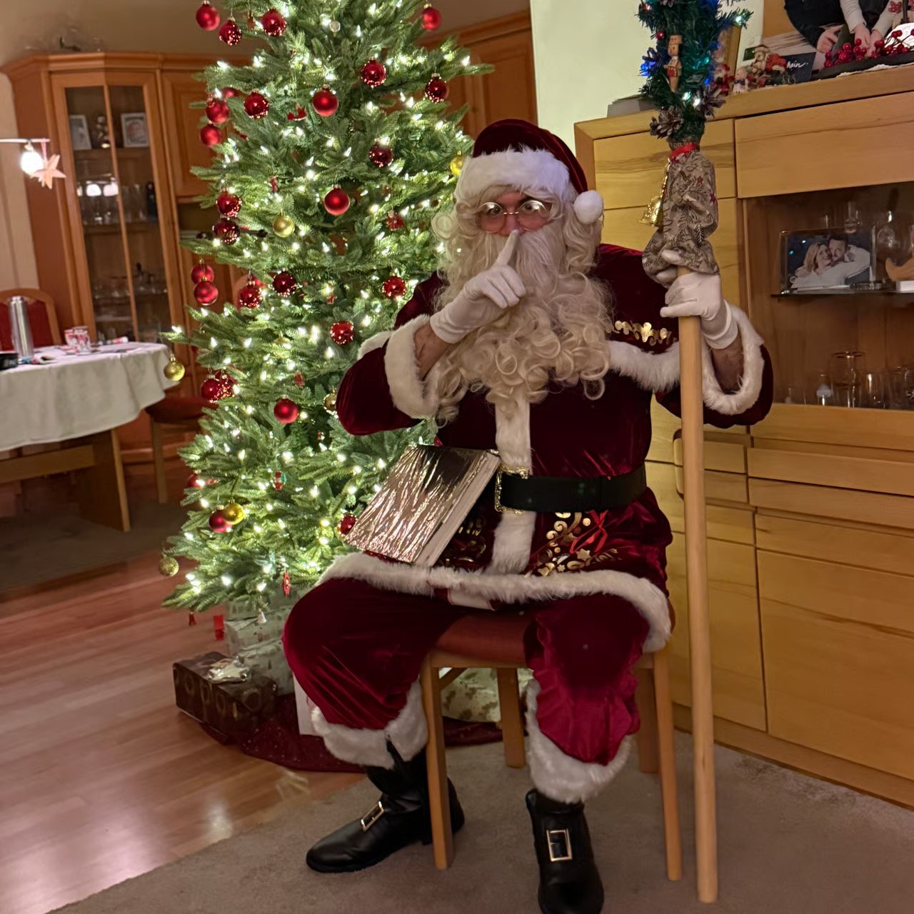 Weihnachtsmann Merlin sitzt mit dem Goldenen Buch vor dem Weihnachtsbaum und hält den Finger vor den Mund